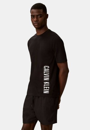 Giovane uomo con maglietta nera Calvin Klein e pantaloncini neri in piedi davanti a uno sfondo semplice con le braccia dietro la schiena.