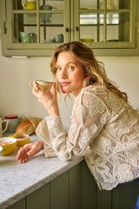 Woman in a cream lace top leaning on a marble kitchen counter, holding a teacup near her face with bowls and bread behind her.