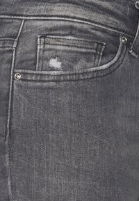 Jeans de mezclilla gris con una textura desvaída, detalle en el bolsillo trasero y acentos de metal. Destacan por su sutil desgaste en la tela.