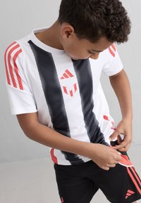 Vit sporttröja med svarta ränder och röda detaljer, med en Adidas-logga. Svarta shorts med matchande röda inslag. Barn som justerar kläderna.