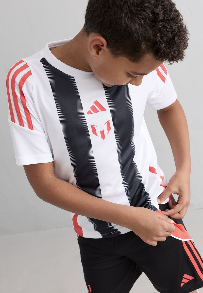 Vit sporttröja med svarta ränder och röda detaljer, med en Adidas-logga. Svarta shorts med matchande röda inslag. Barn som justerar kläderna.