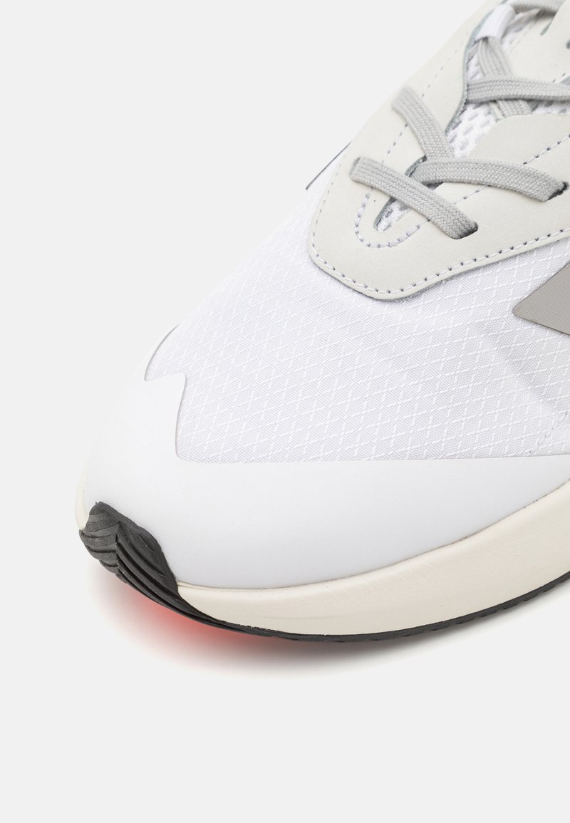 Sneaker atletico bianco con una parte superiore testurizzata, dettagli grigi e suola in gomma nera. Presenta un design con lacci e punta arrotondata.