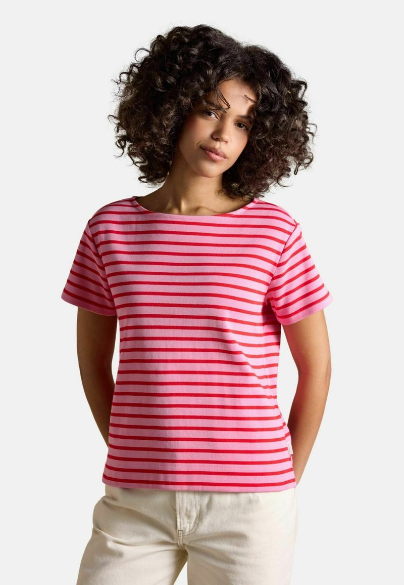 Mujer con cabello rizado, vestida con una camiseta de manga corta a rayas rosas y rojas y pantalones blancos, de pie con las manos detrás de la espalda.