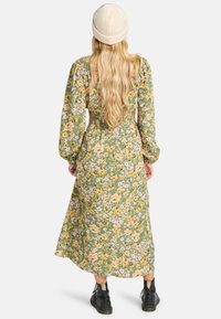 Vestido midi floral en verde, con flores amarillas y moradas, mangas largas y vaporosas, cintura ajustada y un dobladillo fluido. Se lleva con un sombrero de punto beige.