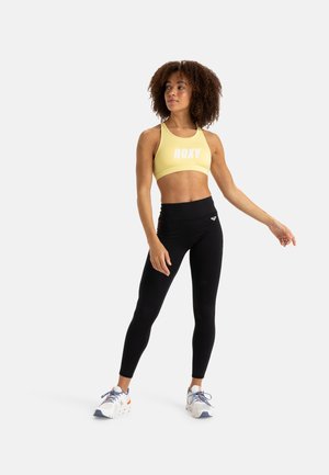 Top sportivo giallo con logo "ROXY", abbinato a leggings neri a vita alta con accenti sottili, indossato con scarpe da ginnastica bianche.