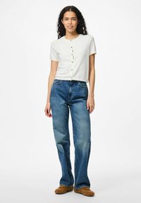 Vit kortärmad knäppt cardigan, kombinerad med blåa löst sittande jeans och bruna skor. En enkel, ren design med funktionella knappar.