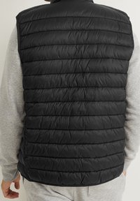 Czarna pikowana kamizelka z poziomym przeszyciem, lekki materiał i wysoki kołnierz; zestawiona z szarą koszulą z długim rękawem pod spodem.