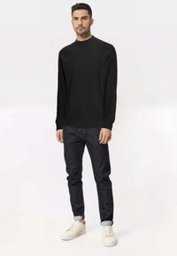 Maglione nero a maniche lunghe con collo alto, abbinato a jeans blu scuro e sneakers bianche, in piedi su uno sfondo chiaro.