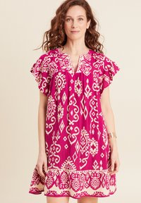 Fuchsia Baumwollkleid mit kurzen Rüschensäumen, das weiße geometrische Muster und einen V-Ausschnitt aufweist. Der ausgestellte Saum sorgt für Bewegung.