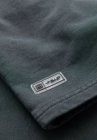 Tela gris oscura con una textura suave. Presenta un logotipo rectangular en negro y plateado que dice "LEVEL UP" en una etiqueta del dobladillo.