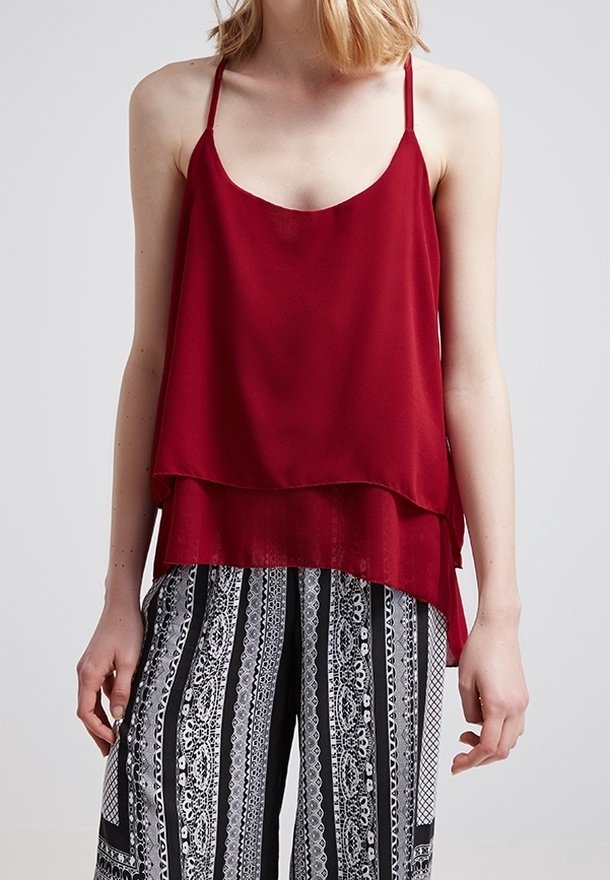 Femme portant un débardeur rouge ample à couches sans manches et un pantalon large à motifs noirs et blancs sur un fond uni.