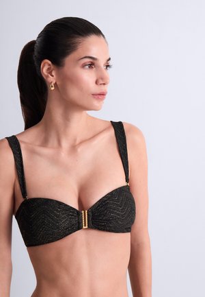 Femme aux cheveux foncés attachés en queue de cheval, portant des boucles d'oreilles en or et un haut de bikini noir texturé avec une fermeture dorée, regardant vers la droite sur un fond uni.