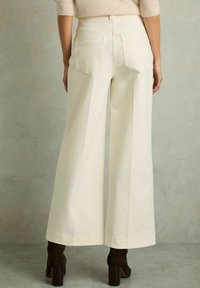 Široké kalhoty off-white barvy z těžkého bavlněného materiálu s jemnou texturou; mají dvě zadní kapsy a vysoký pas. Nošeny s tmavými botami na podpatku.