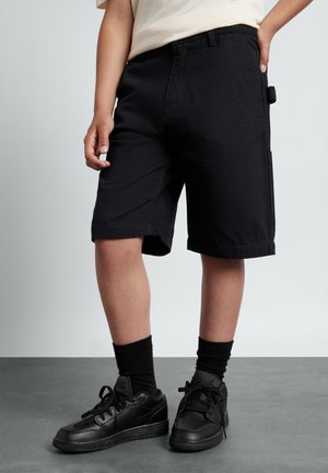 Sorte shorts lavet af et slidstærkt stof med lige snit, udstyret med sidelommer og båret med sorte sneakers og sokker.