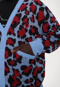 Cardigan bleu avec imprimé léopard rouge et noir audacieux, boutons et bordure bleu clair, porté sur une chemise noire, main partiellement dans la poche.
