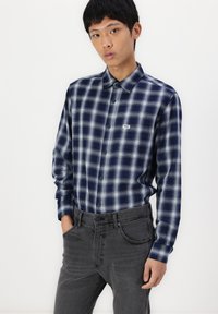 Ung mann iført en blå-hvit rutete knappet skjorte, stukket inn i mørkegrå jeans, står med den ene hånden i lommen.