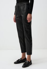Schwarze Kunstlederhosen mit hoher Taille, Falten und schmal zulaufenden Beinen. Kombiniert mit schwarzen Slippern mit Quastenakzenten.