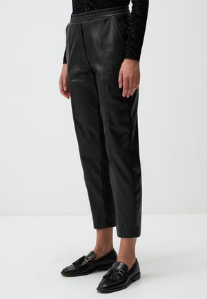 Zwarte faux leren broek met hoge taille, plooien en taps toelopende pijpen. Gecombineerd met zwarte loafers met kwastjesaccenten.