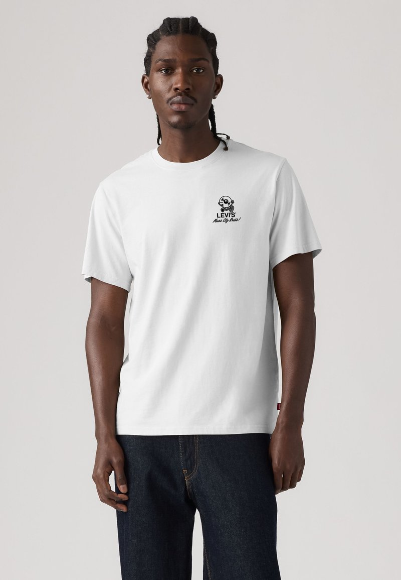Άνδρας με πλεξούδες φοράει λευκό t-shirt Levi's με μικρό γραφικό και κείμενο αριστερά στο στήθος και σκούρο μπλε τζιν.