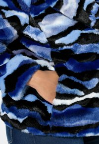 Veste en fausse fourrure présentant un motif ondulé dans des nuances de bleu, noir et blanc ; texture douce avec un long poil, et un détail de poche latérale.