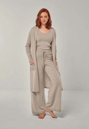 Woman with red hair standing barefoot wearing beige knit tank top, wide-leg pants, and long cardigan with pockets on a neutral background.