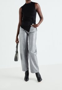 Top sans manches noir avec un détail noué, associé à un pantalon large argenté brillant et des bottines noires à bout pointu, portant un sac à bandoulière gris.