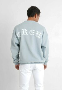 Felpa blu chiaro con collo a giro e lettering bianco testurizzato "CREW" sulla schiena. Presenta una vestibilità rilassata e maniche lunghe. Abbinata a pantaloni bianchi.