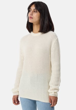 Cremefarbener gestrickter Pullover mit langen Ärmeln, gerippter Textur und lässiger Passform. Rundhalsausschnitt und Tropfensaum, kombiniert mit blauen Jeans.