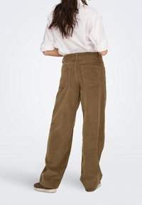 Femme portant un pantalon large en velours côtelé marron, une chemise blanche aux manches retroussées et des baskets marron, debout, les mains sur les hanches, tournée dans le dos.