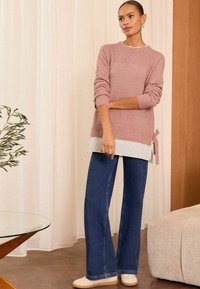 Svijetloružičasti pleteni pulover s čipkastim uzorkom, složen preko bijele majice. Uparen s širokim plavim trapericama i bež cipelama. U blizini se nalazi drveni i stakleni stol.