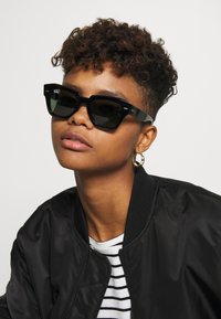 Ochelari de soare negri, rectangulare, cu rame groase, lentile întunecate și un branding subtil pe lateral, asortați cu o geacă bomber neagră și o cămașă cu dungi.