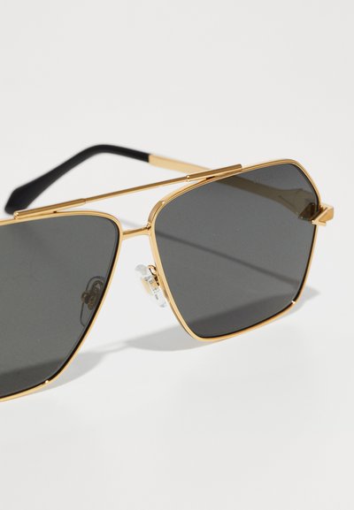 Slnečné okuliare typu aviator v zlatom ráme s tmavými tónovanými šošovkami a čiernymi koncami straníc na bielom povrchu.