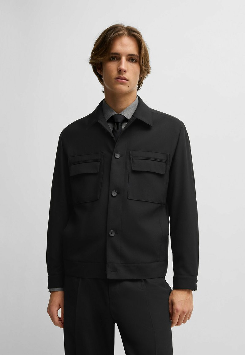 Giacca nera con bottoni e due tasche sul petto, texture liscia e design con colletto. Abbinata a una camicia grigia e una cravatta.