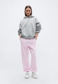 Γυναίκα που στέκεται με τα χέρια στις τσέπες, φορώντας γκρι φαρδιά φούτερ, ροζ φόρμες, λευκά αθλητικά παπούτσια και ασημένια σκουλαρίκια σε σχήμα κρίκου σε λευκό φόντο.