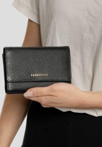 Cartera de cuero negro con un acabado texturizado, que presenta una solapa frontal y un sutil logo dorado "LAZAROTTI". Sostenida con una mano.