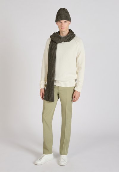 Young man wearing a white sweater, olive green pants, white sneakers, and matching dark green knit hat and long scarf against plain background.