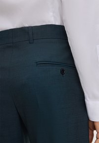 Close-up van een persoon in een donkergroene, op maat gemaakte broek met een knoopzak aan de achterkant en een wit overhemd met lange mouwen.