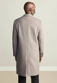 Beżowy wełniany płaszcz z pojedynczym rozcięciem z tyłu, dopasowany krój i zapięcie na guziki przy mankietach. Gładka faktura i pełna długość.