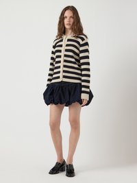 Cardigan a righe in crema e navy, con chiusura a bottoni e maniche lunghe. Abbinato a una gonna scura con volant e scarpe nere.