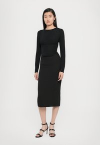 Zwarte lange mouwen top met een aansluitend lijfje, gecombineerd met een hoge zwarte kokerrok. Model draagt zwarte bandjeshakken. Glad textuur overal.