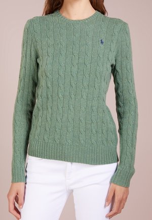 Femme vêtue d'un pull vert à torsades à manches longues et d'un pantalon blanc, debout devant un fond uni.
