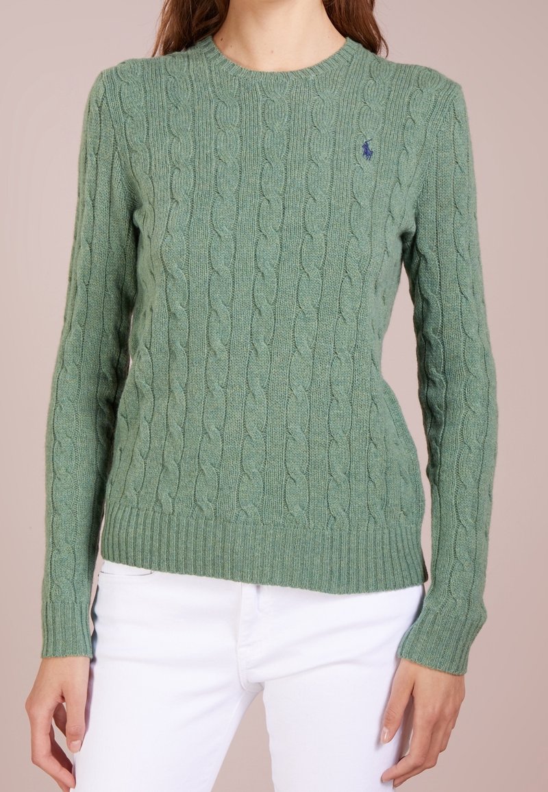 Femme vêtue d'un pull vert à torsades à manches longues et d'un pantalon blanc, debout devant un fond uni.