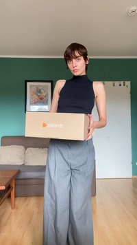 Barna Zalando doboz egy személy előtt, aki fekete ujjatlan toppot és szürke, bőszárú nadrágot visel, a háttérben zöld fallal.