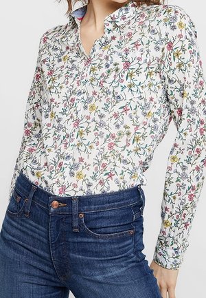 Mujer que lleva una camisa blanca abotonada con un patrón floral colorido metida en unos vaqueros azul oscuro de tiro alto.