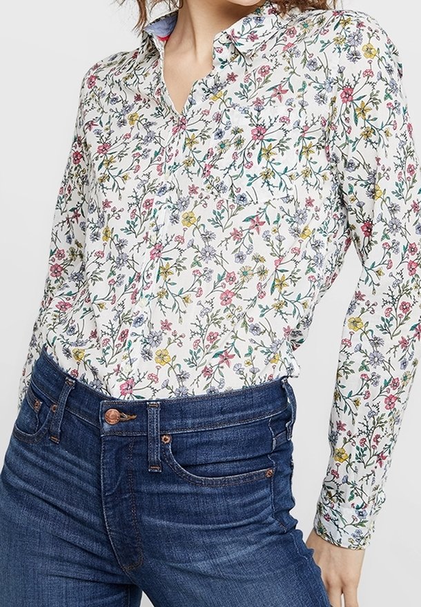 Femme portant une chemise blanche à boutons avec un motif floral coloré, rentrée dans un jean bleu foncé taille haute.