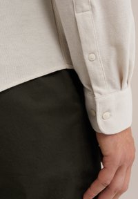 Krém színű, texturált felületű ing hosszú ujjakkal, gombos mandzsettával, és zöld olajszínű derekú részt láthatunk alul.