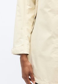 Μπεζ αδιάβροχο μπουφάν με απαλή υφή, κουμπιά snap και λογότυπο στο μανίκι. Η μανσέτα είναι διπλωμένη για ρυθμιζόμενη εφαρμογή.