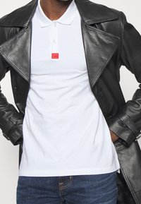 Weißes Polohemd mit kurzem Kragen und rotem "HUGO"-Label, unter einer schwarzen Lederjacke mit breiten Revers getragen. Dunkelblaue Jeans sichtbar.