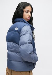 Hosszú fekete hajú nő kék The North Face pufi dzsekiben, vállán túlra nézve egy sima világos háttér előtt.