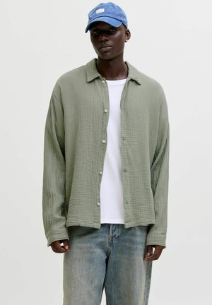 Jeune homme portant une chemise à boutons vert clair sur un t-shirt blanc, un jean délavé ample, et une casquette de baseball bleue, debout devant un fond uni.